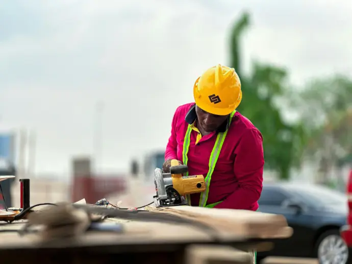 A worker cutting down wooden boards at a construction site, he is wearing yellow safety hat